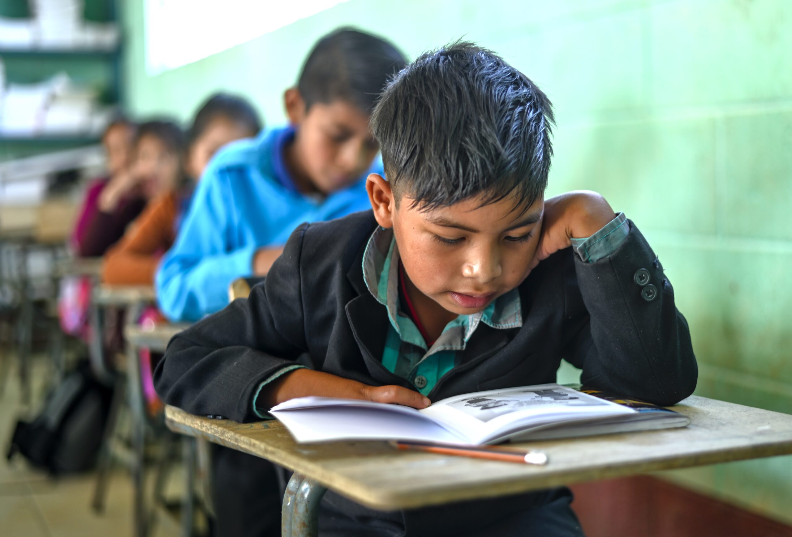 Young Guatemalan boy at a school desk looking down and reading a book.