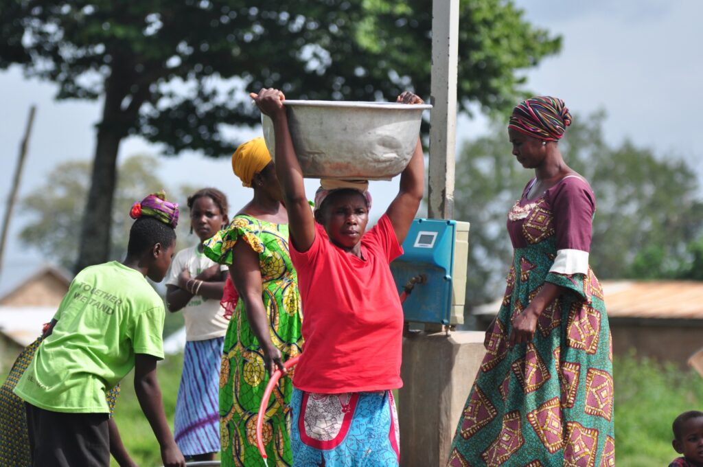 Primary focus of a woman in Ghana transporting a large pot of water on her head with people in the background near the water system