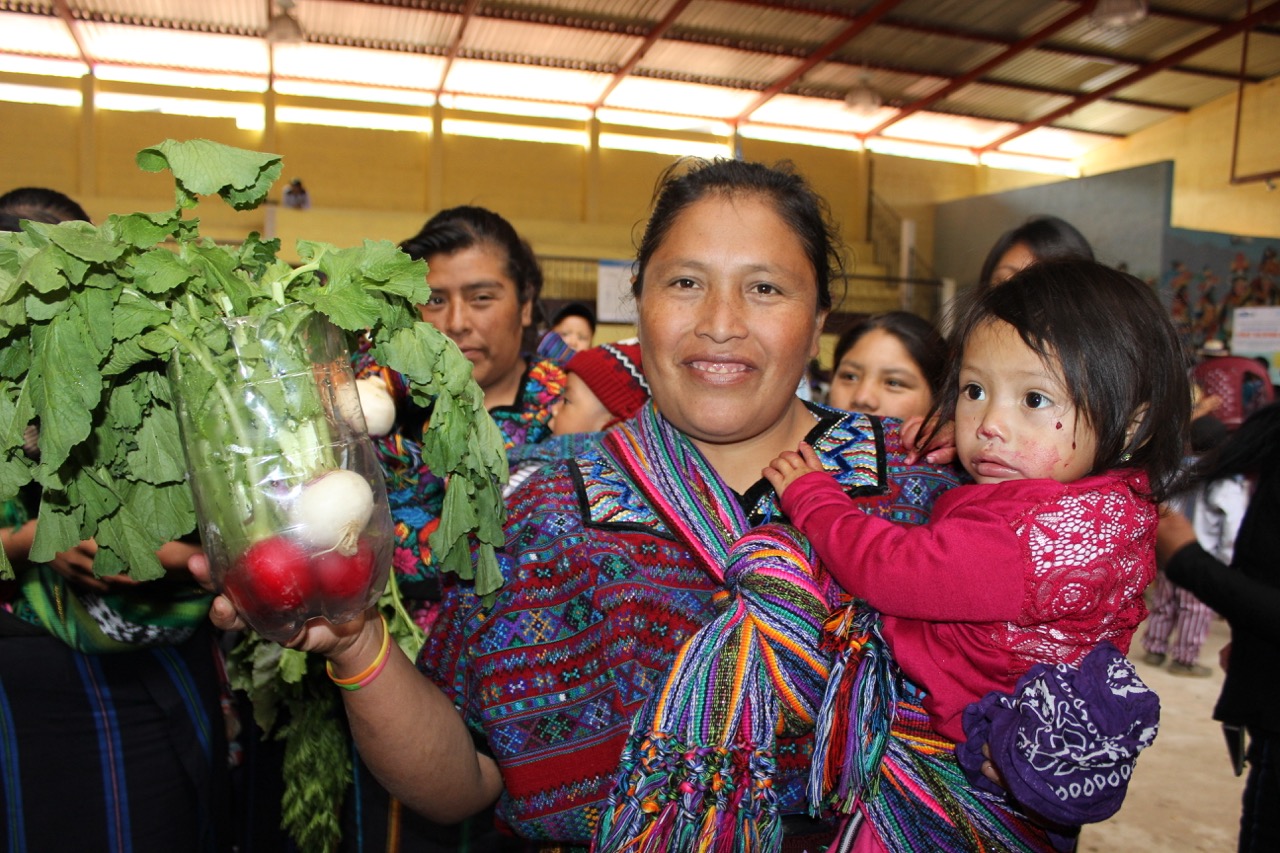 A woman carrying her child in a sling holding fresh vegetables in her free hand and smiling at the camera.